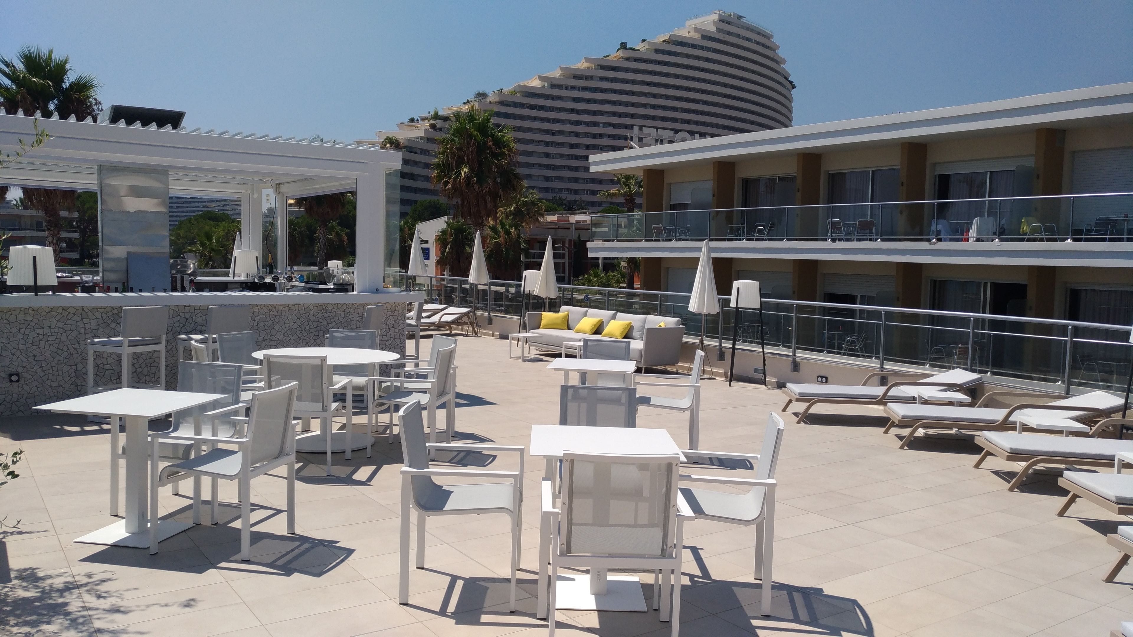 A rooftop bar with white tables and chairs, a white sofa with yellow cushions, and a poolside area with lounge chairs.