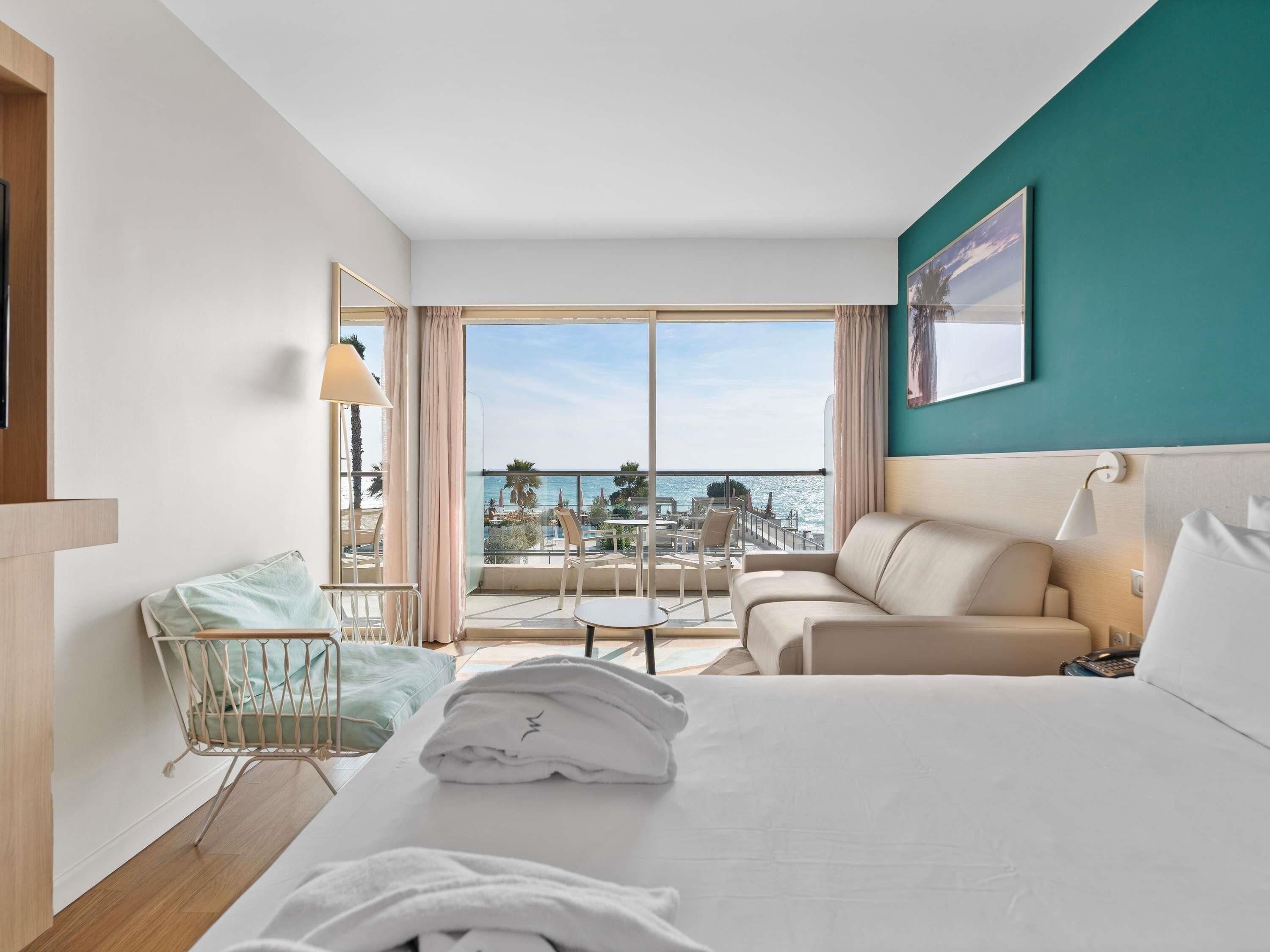 A hotel room with a bed, a sofa, a small table, a chair, a balcony with a view of the sea, and a painting on the wall.