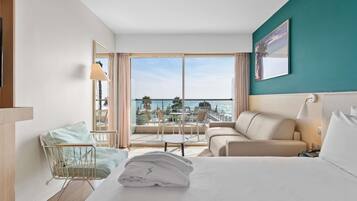 Quarto de hotel com cama, sofá, mesinha, cadeira, varanda com vista para o mar e um quadro na parede.