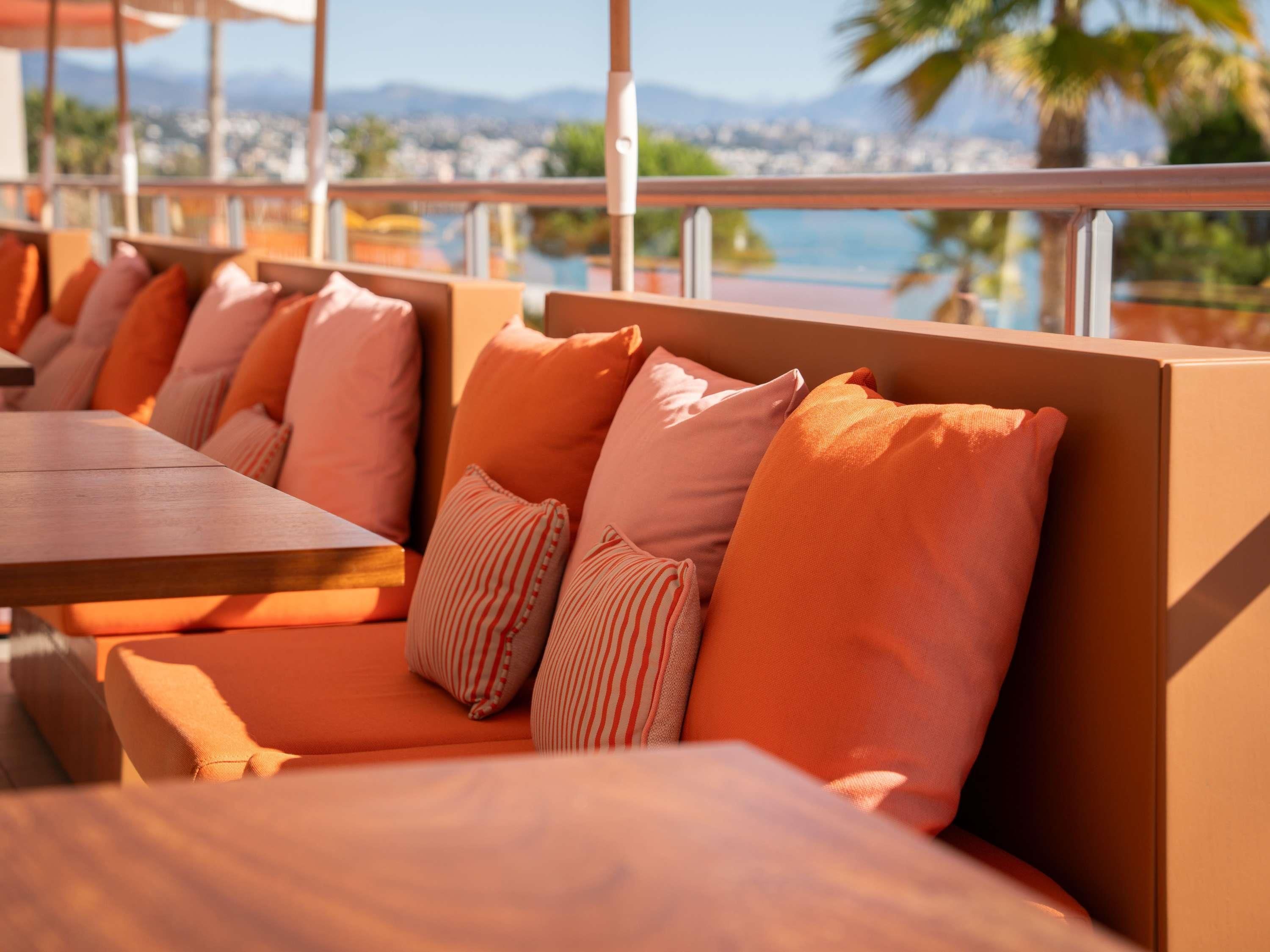 Una fila de asientos tapizados en color naranja con almohadones a rayas, una mesa de madera y vistas a un paisaje urbano y montañas a través de amplios ventanales.
