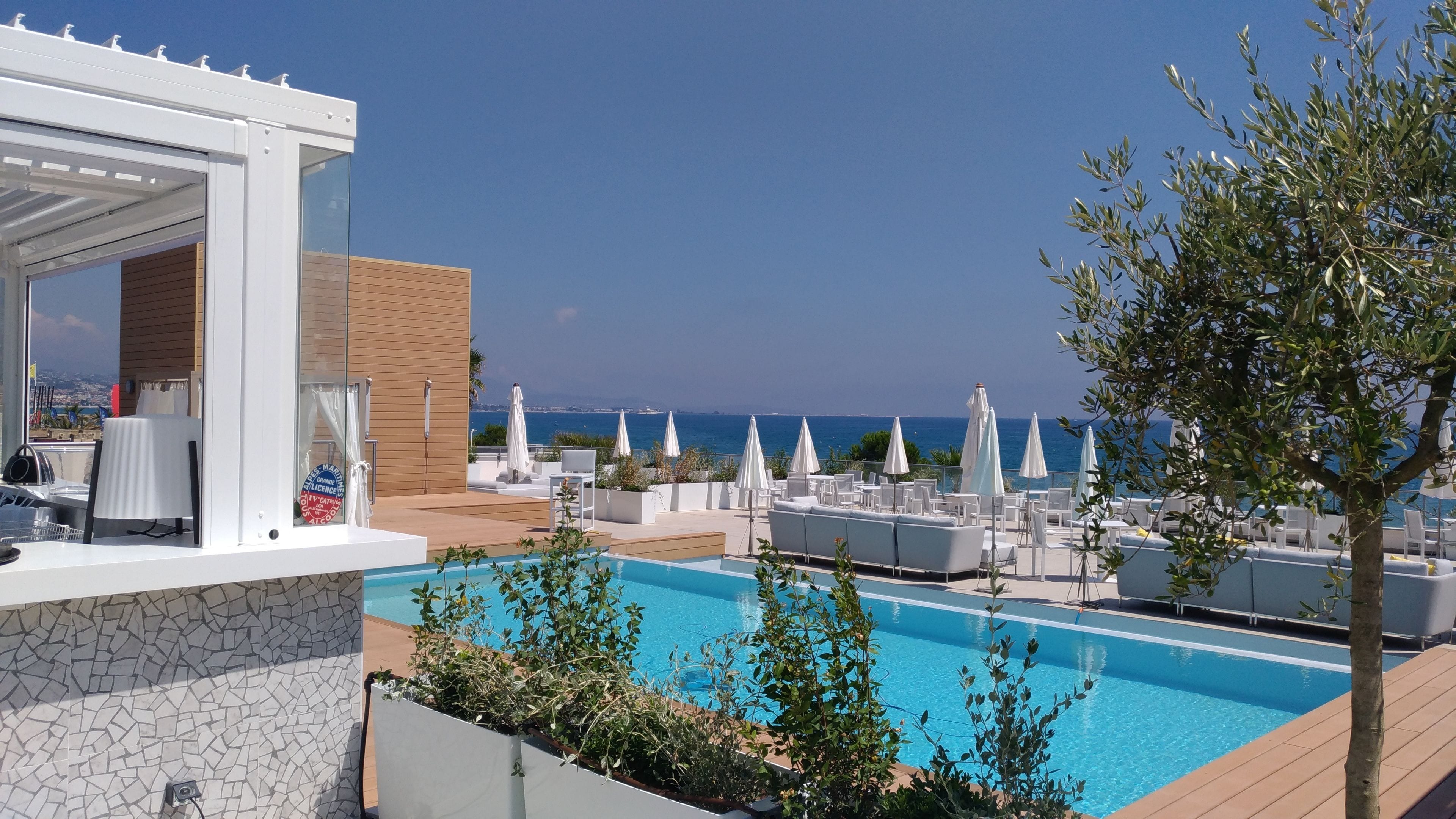 A rooftop pool area with lounge chairs and umbrellas, overlooking a clear blue sea.
