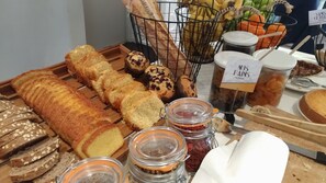 Variedad de panes y pasteles expuestos sobre una tabla de madera, con frascos de mermeladas y una cesta de frutas al fondo.
