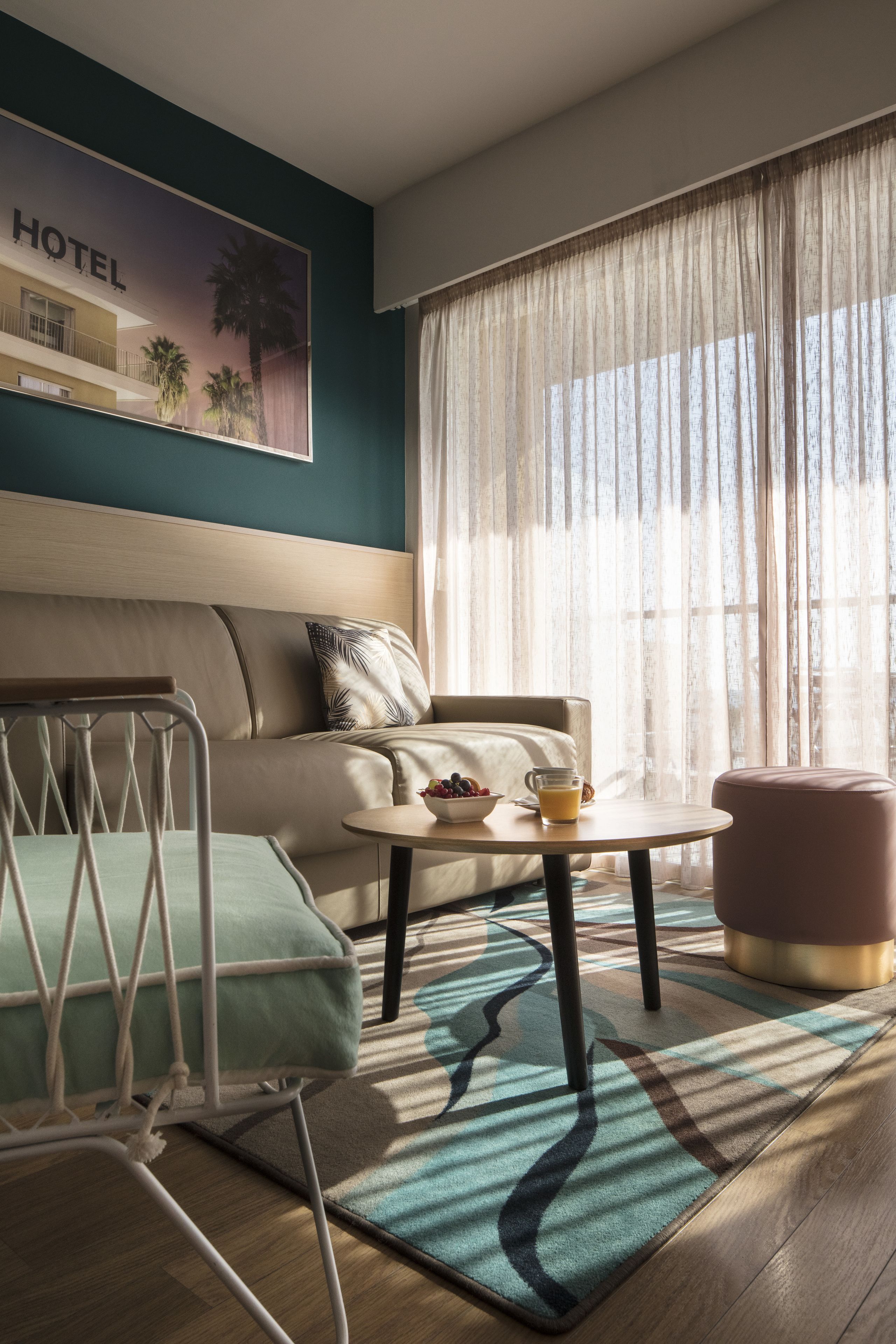 A hotel room with a sofa, a small table with a bowl of fruit and a glass of orange juice, a chair, and a window with sheer curtains.