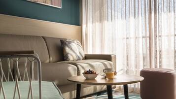 A hotel room with a sofa, a small table with a bowl of fruit and a glass of orange juice, a chair, and a window with sheer curtains.
