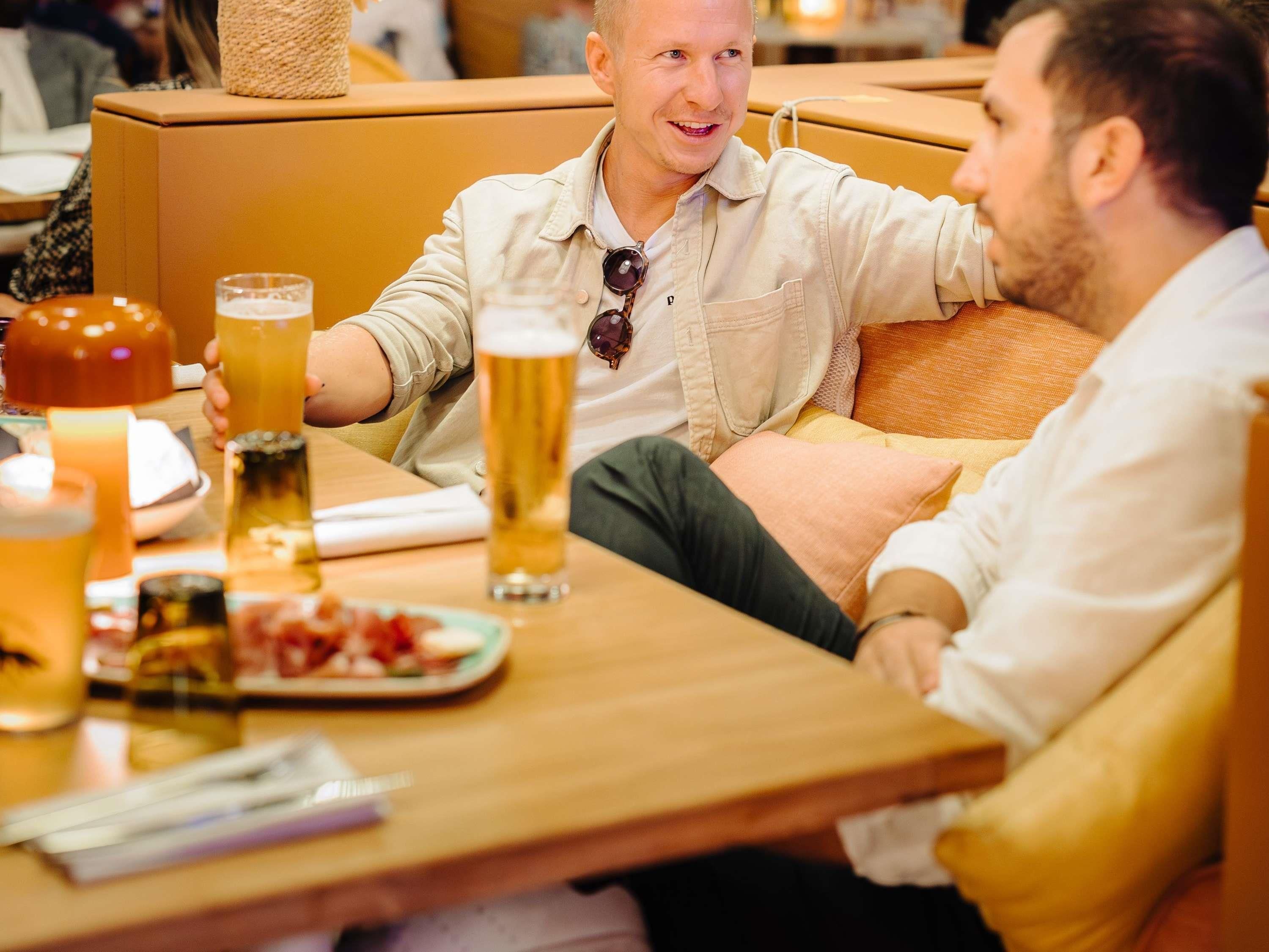 Two people sitting at a table with drinks and food, in a room with a couch and a wooden table.