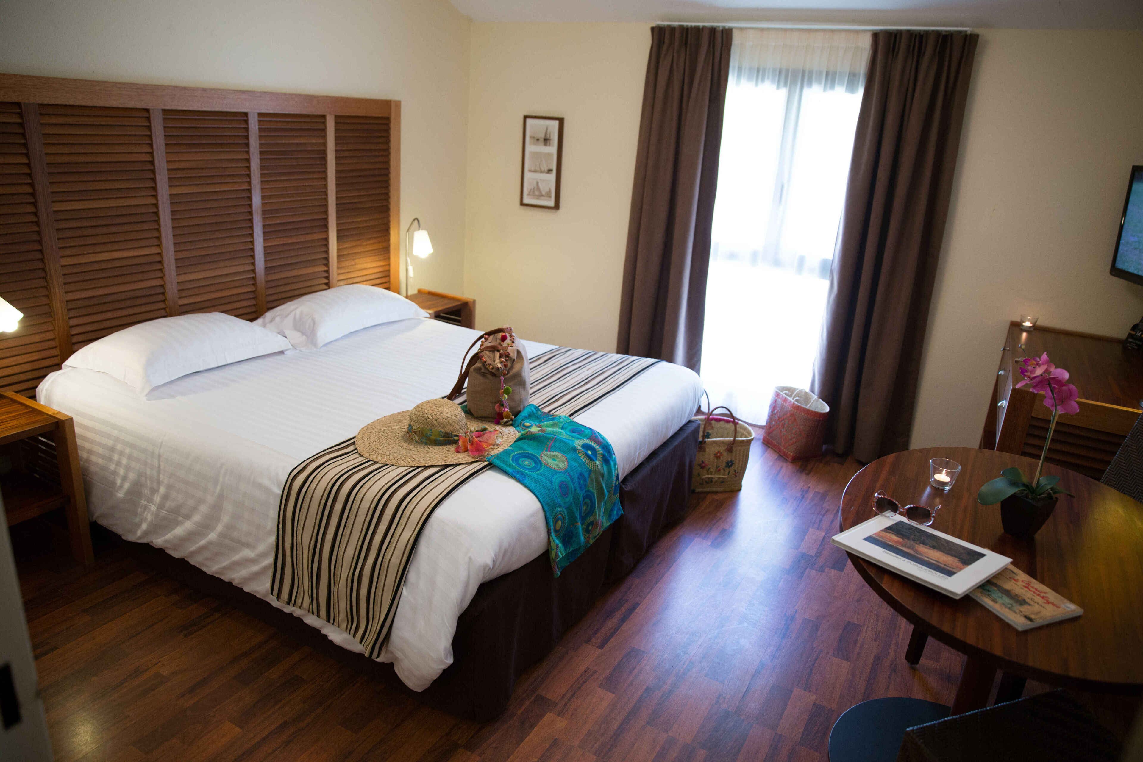 Quarto de hotel com uma cama grande, cabeceira de madeira, mesas de cabeceira com luminárias, uma pequena mesa com um vaso de flores e uma cadeira.