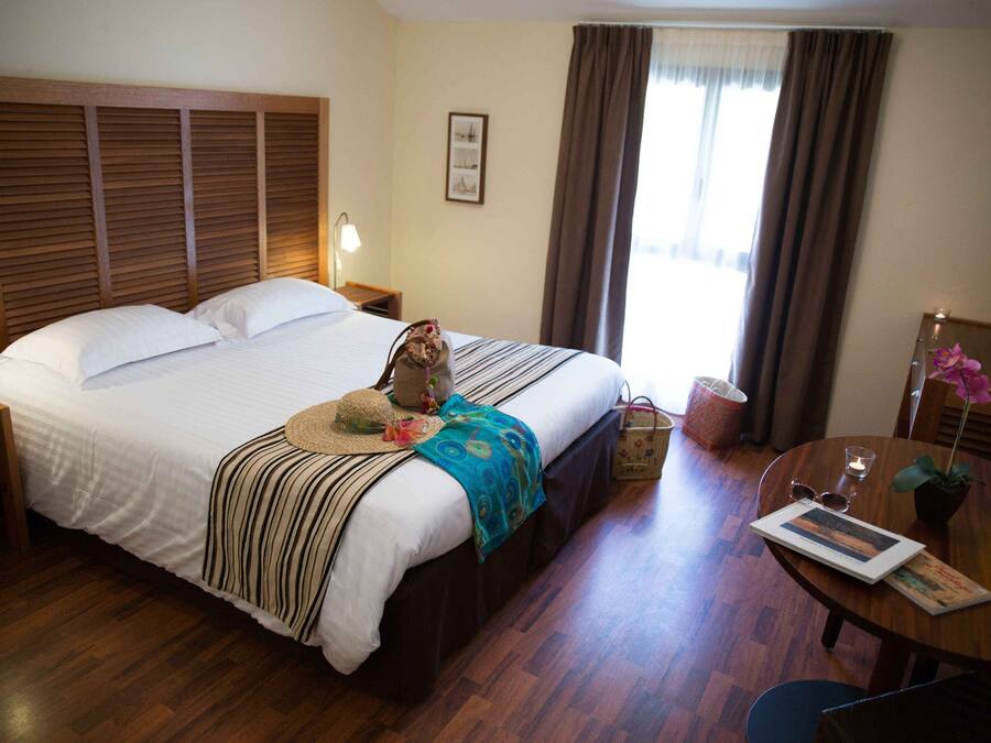 Quarto de hotel com uma cama grande, cabeceira de madeira, mesas de cabeceira com luminárias, uma pequena mesa com um vaso de flores e uma cadeira.
