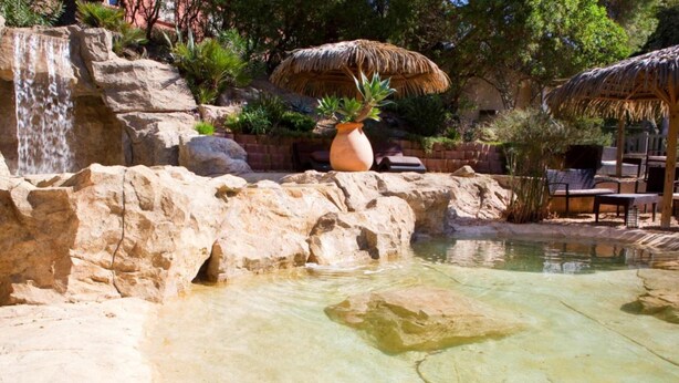 Une formation rocheuse avec un petit bassin, un grand pot en terre cuite et des parasols en chaume.