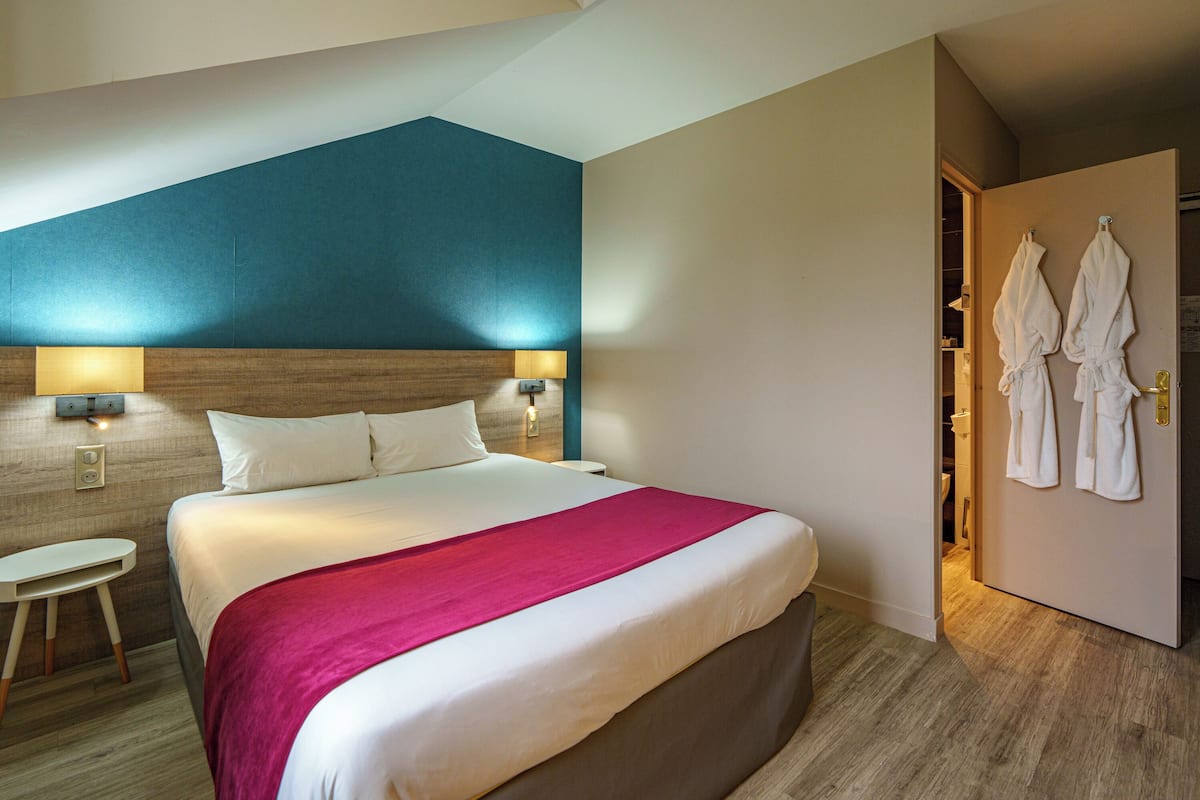 Une chambre d’hôtel avec un grand lit, deux lampes de chevet, une tête de lit en bois, un plaid rouge et une porte sur laquelle sont accrochés deux peignoirs blancs.