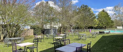 Un patio al aire libre con mesas y sillas, rodeado de árboles y una piscina al fondo.