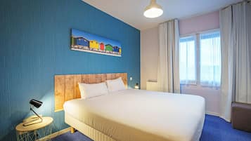 Ein Hotelzimmer mit einem großen Bett, einem Holz-Kopfteil, einem Gemälde an der Wand, einem runden Beistelltisch mit Lampe und einem Fenster mit Vorhängen.