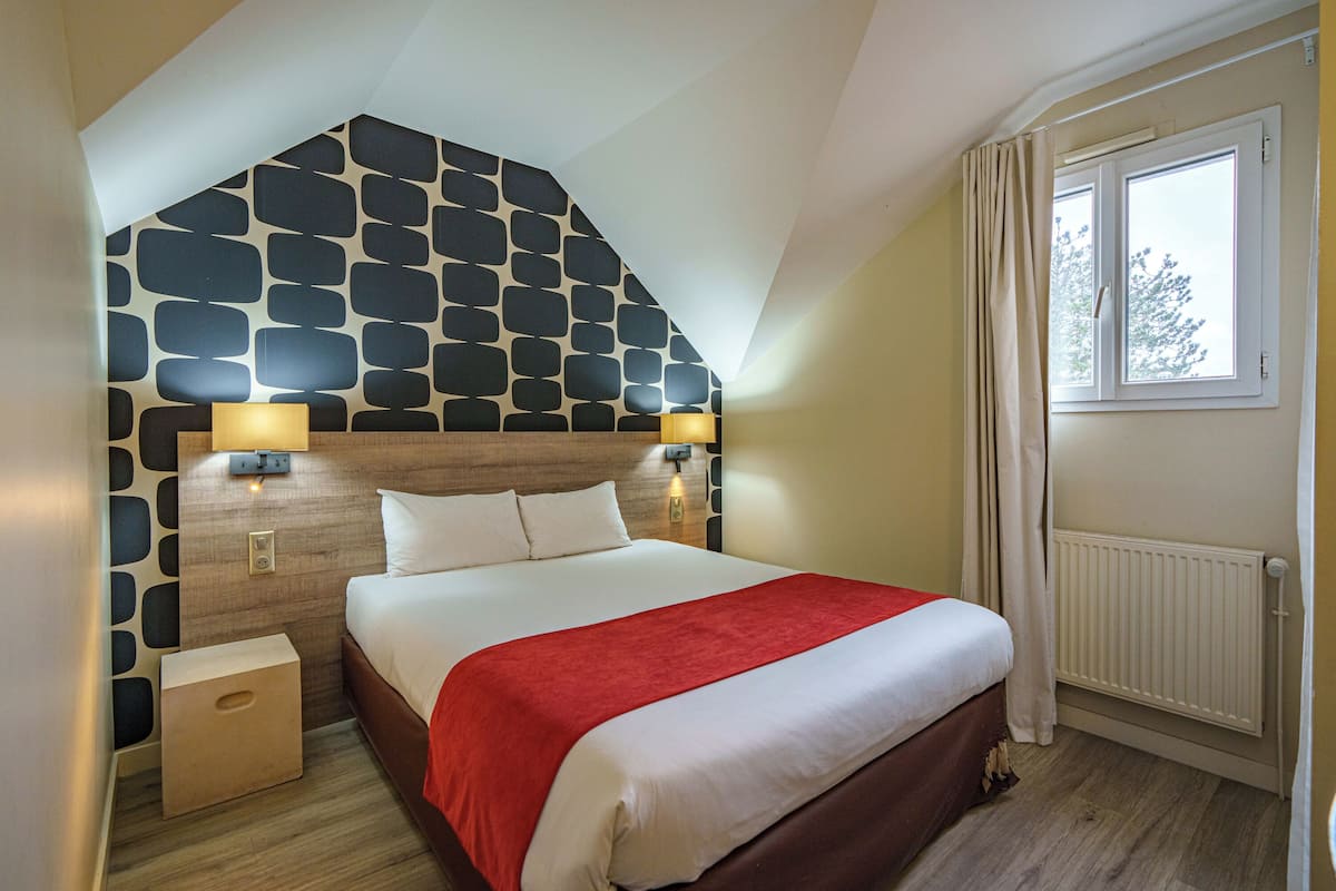 Une chambre avec un plafond en pente, un lit recouvert d’une couverture rouge, deux lampes de chevet, une tête de lit en bois et une fenêtre avec des rideaux.