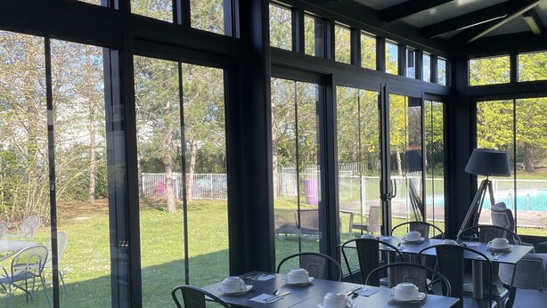 Une véranda avec des tables dressées pour le repas, de grandes fenêtres et une vue sur l’extérieur.
