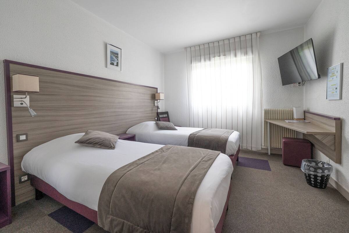 A hotel room with two beds, a desk, a TV, and a window with curtains.