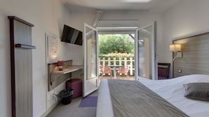 A hotel room with a bed, a desk with a lamp, a TV mounted on the wall, and a view of the outside. - Contact Hôtel O’Nice Saintes (Saintes)