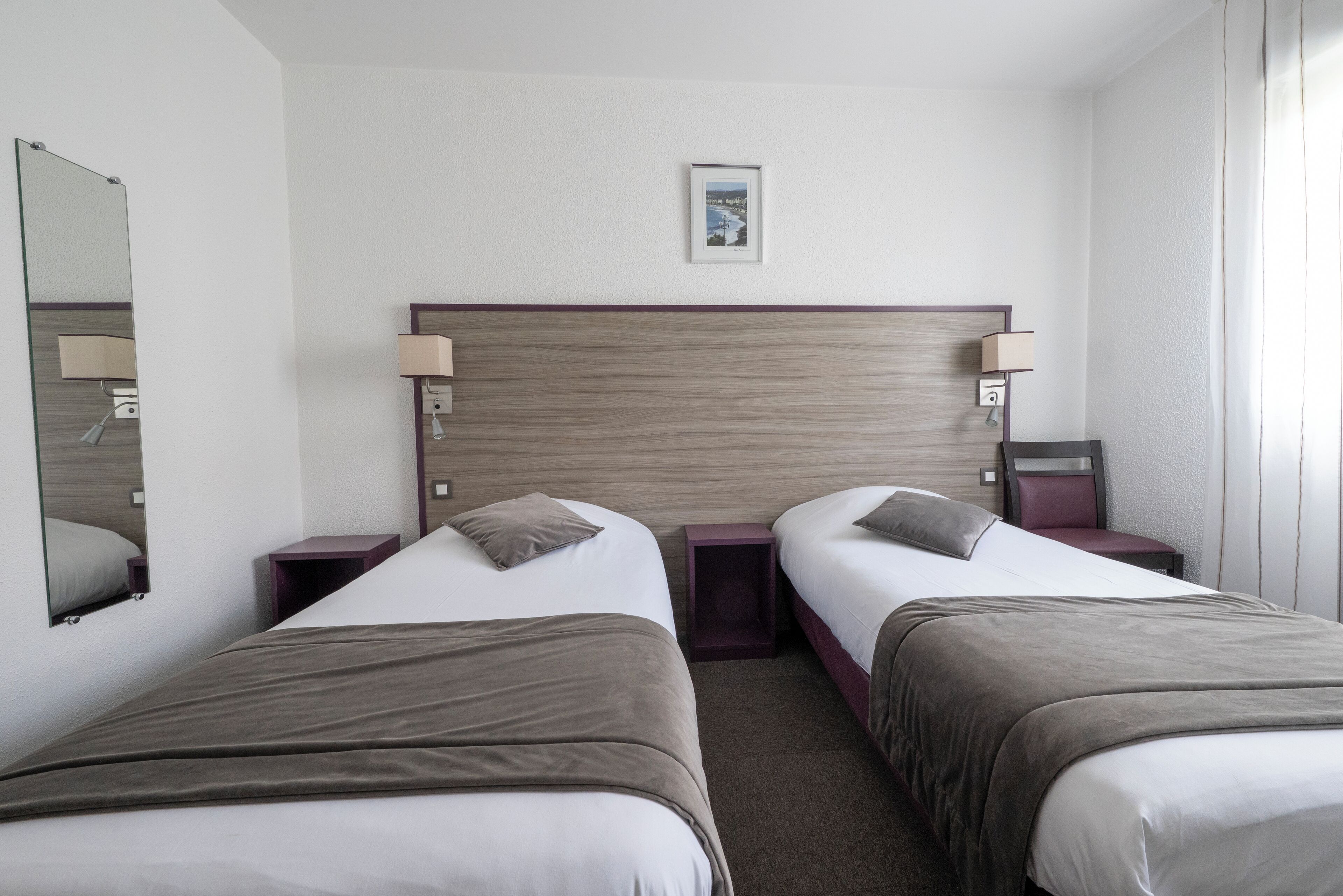 A hotel room with two beds, a wooden headboard, bedside tables with lamps, a framed picture on the wall, and a window with curtains.