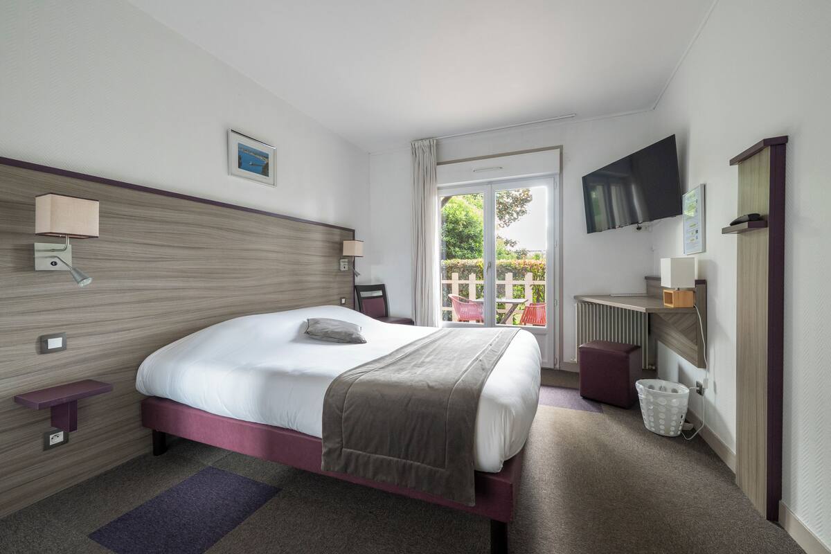 A hotel room with a bed, bedside lamps, a TV, a small table, a chair, and a view of the outdoors.
