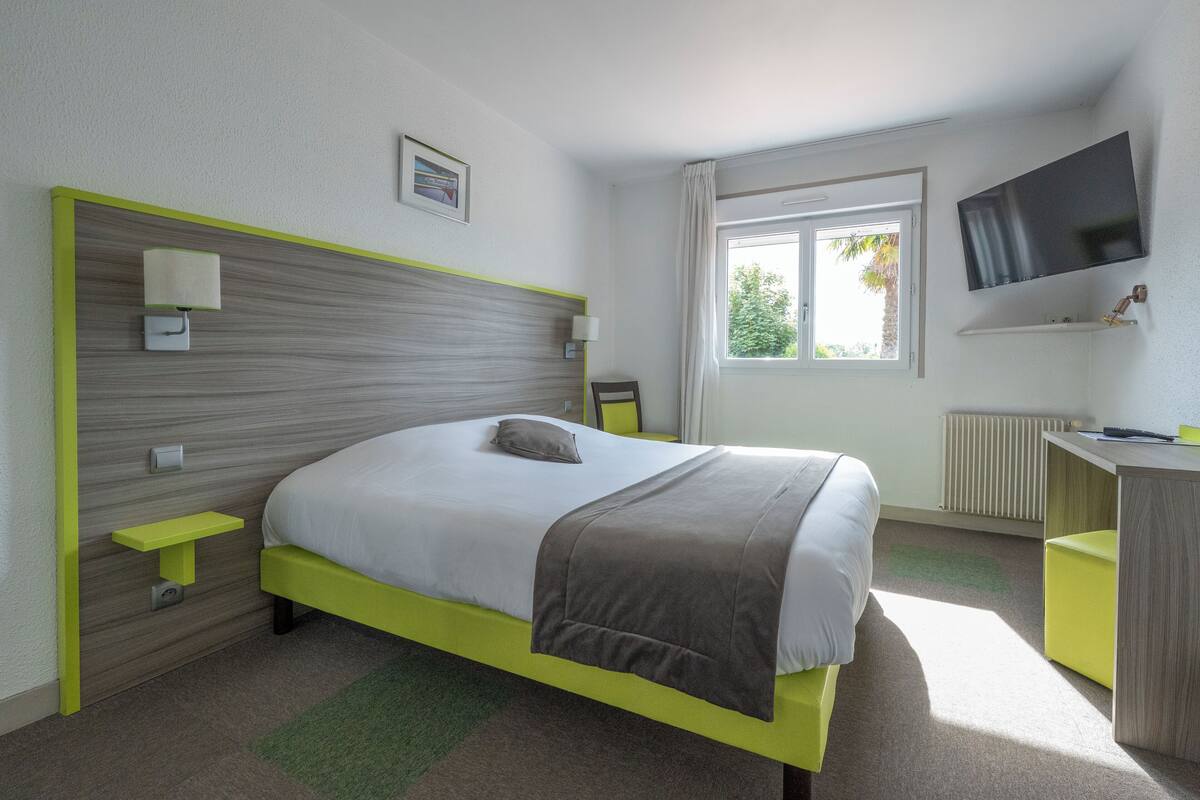 A modern hotel room with a bed, bedside tables, a desk, a TV, and a window with a view of trees.