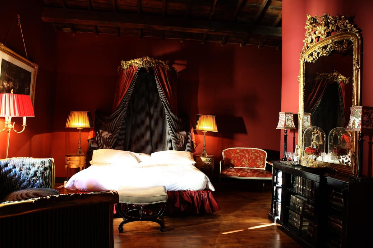 A room with a large bed, two bedside tables with lamps, a red armchair, a wooden cabinet, and a large ornate mirror.