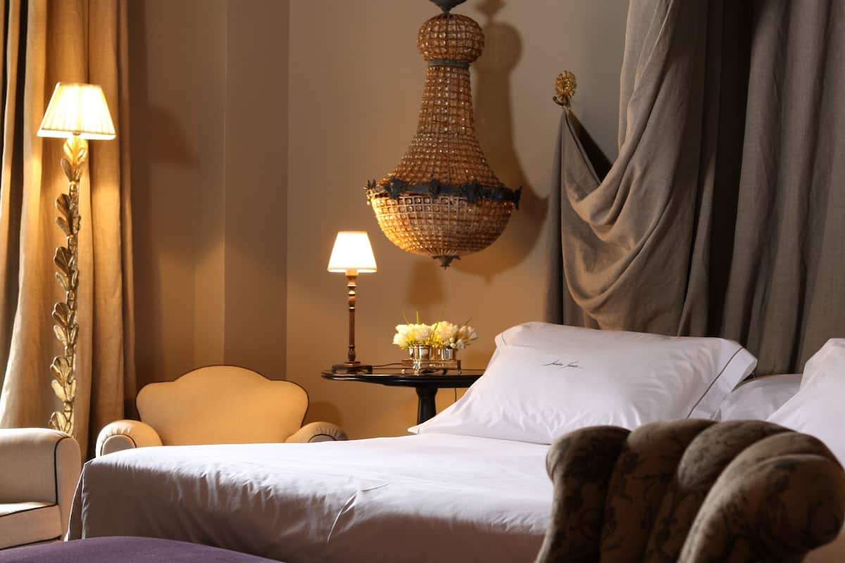A hotel room with a bed, a chair, a nightstand with lamps, and a hanging chandelier.