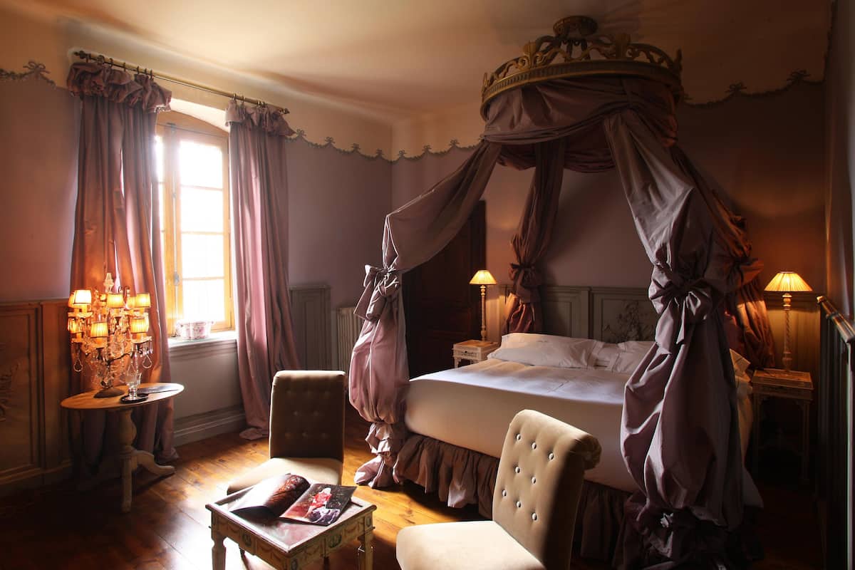 A bedroom with a four-poster canopy bed, a chandelier, and a table with a magazine.