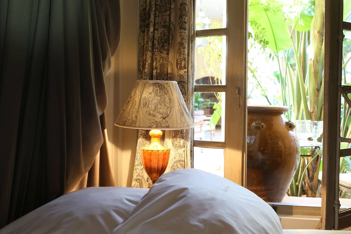 A bed with white linens, a lamp with a patterned shade, and a view of greenery through an open door.