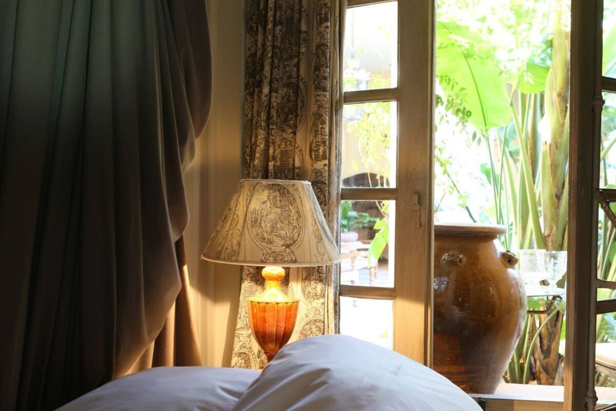 A bed with white bedding, a lamp with a patterned lampshade, and a view of greenery through an open window.