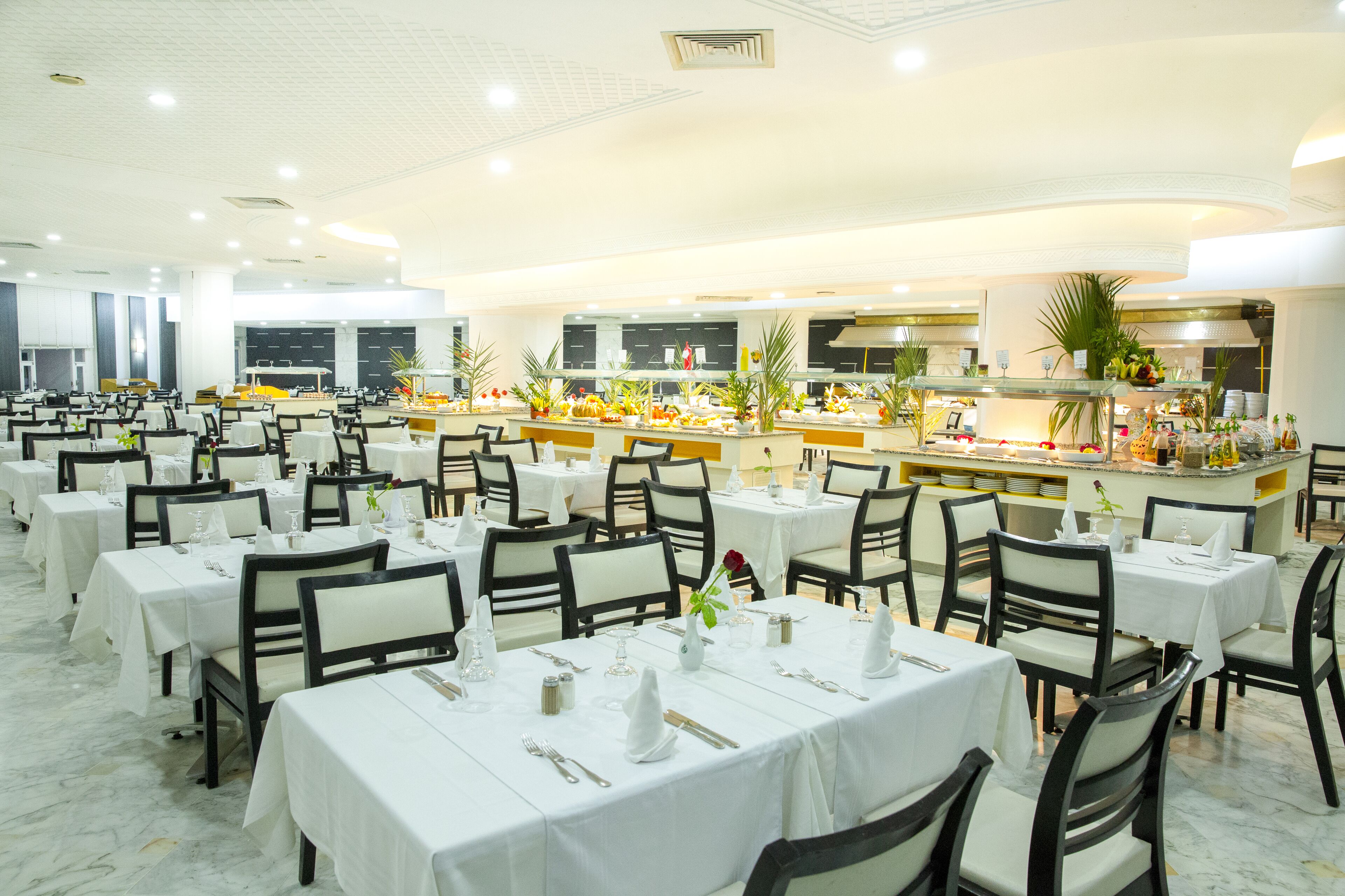 Un'ampia sala da pranzo con numerosi tavoli e sedie, un buffet centrale ricco di cibi diversi e una cucina moderna sullo sfondo.