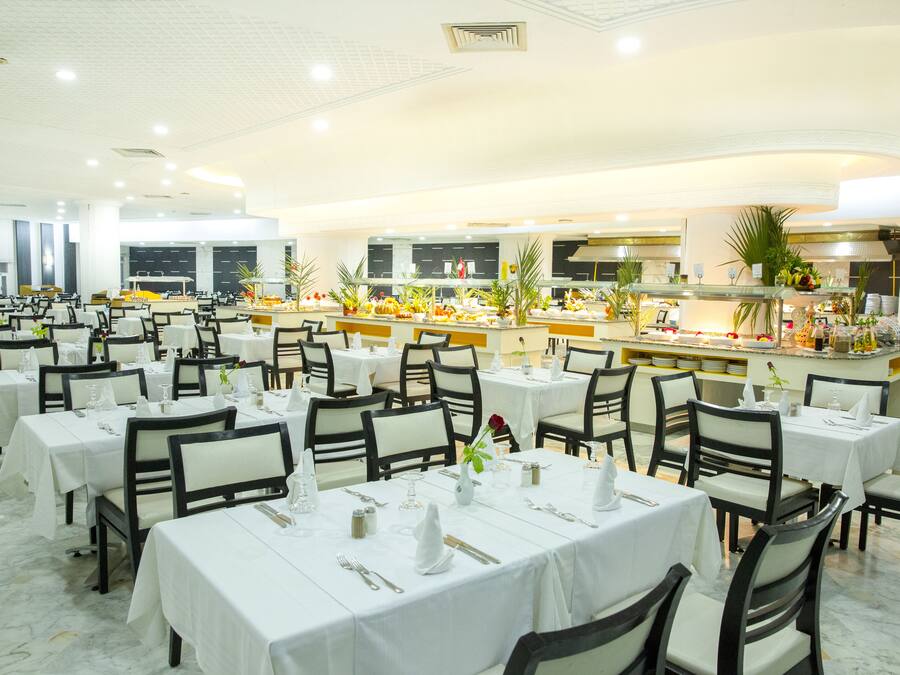 Un'ampia sala da pranzo con numerosi tavoli e sedie, un buffet centrale ricco di cibi diversi e una cucina moderna sullo sfondo.