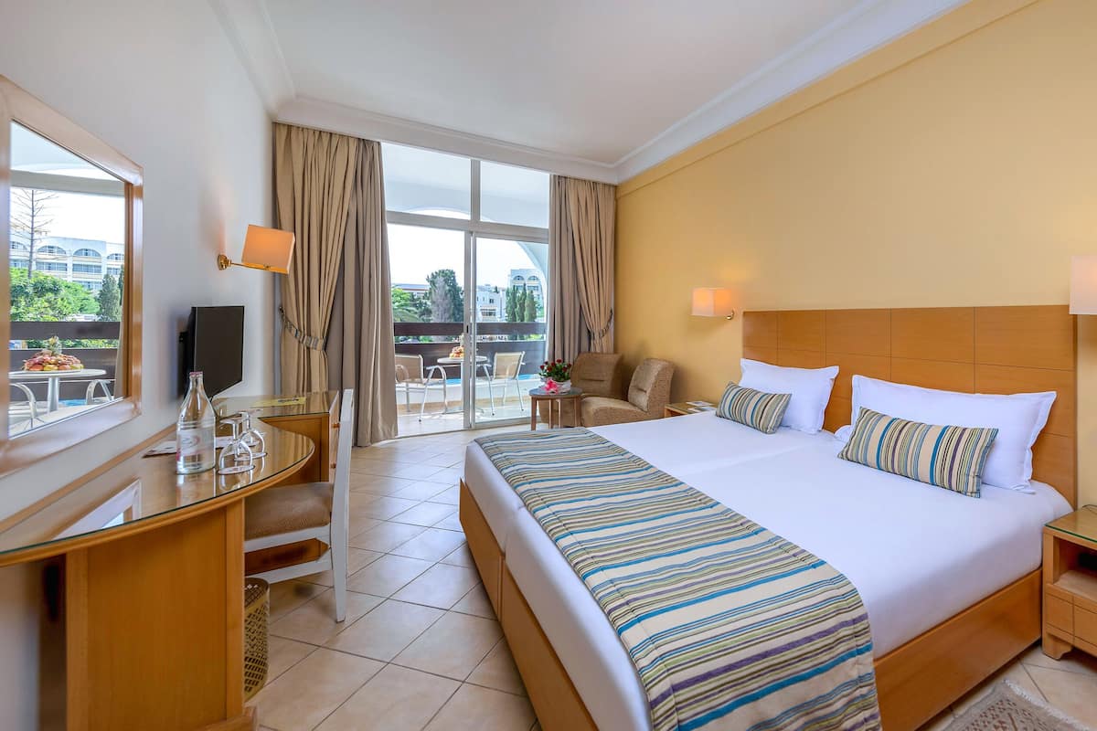 Habitación de hotel con una cama grande, un escritorio con silla, televisión y un balcón con mobiliario exterior.