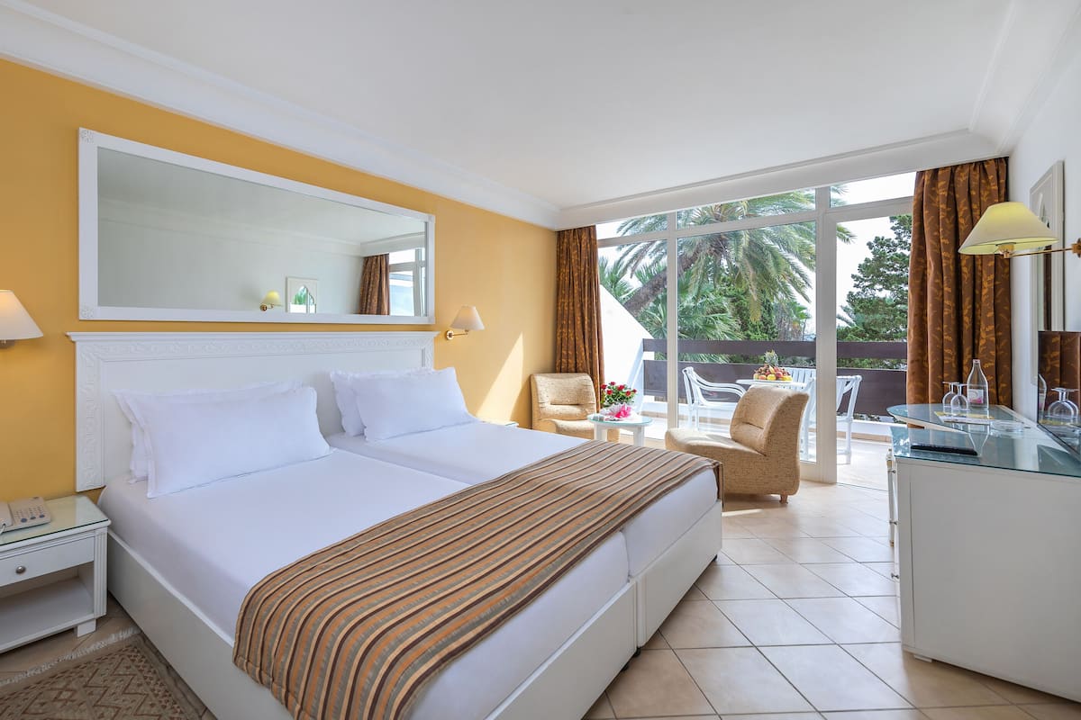 Habitación de hotel con una cama grande, un escritorio con lámpara, una silla y un balcón con mesa y sillas.