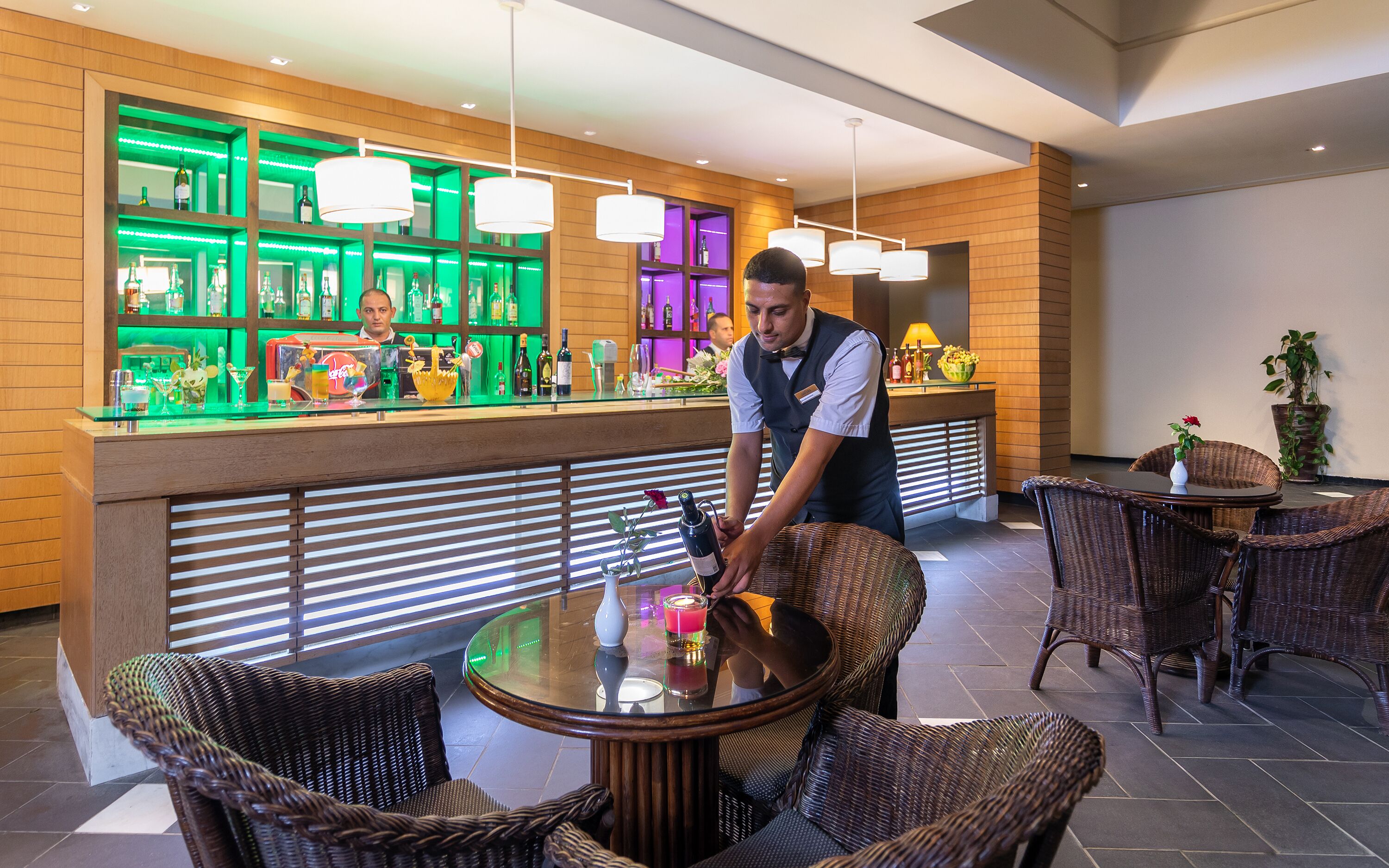 Uma área de bar com um bartender, móveis de vime e uma prateleira iluminada em verde com garrafas.