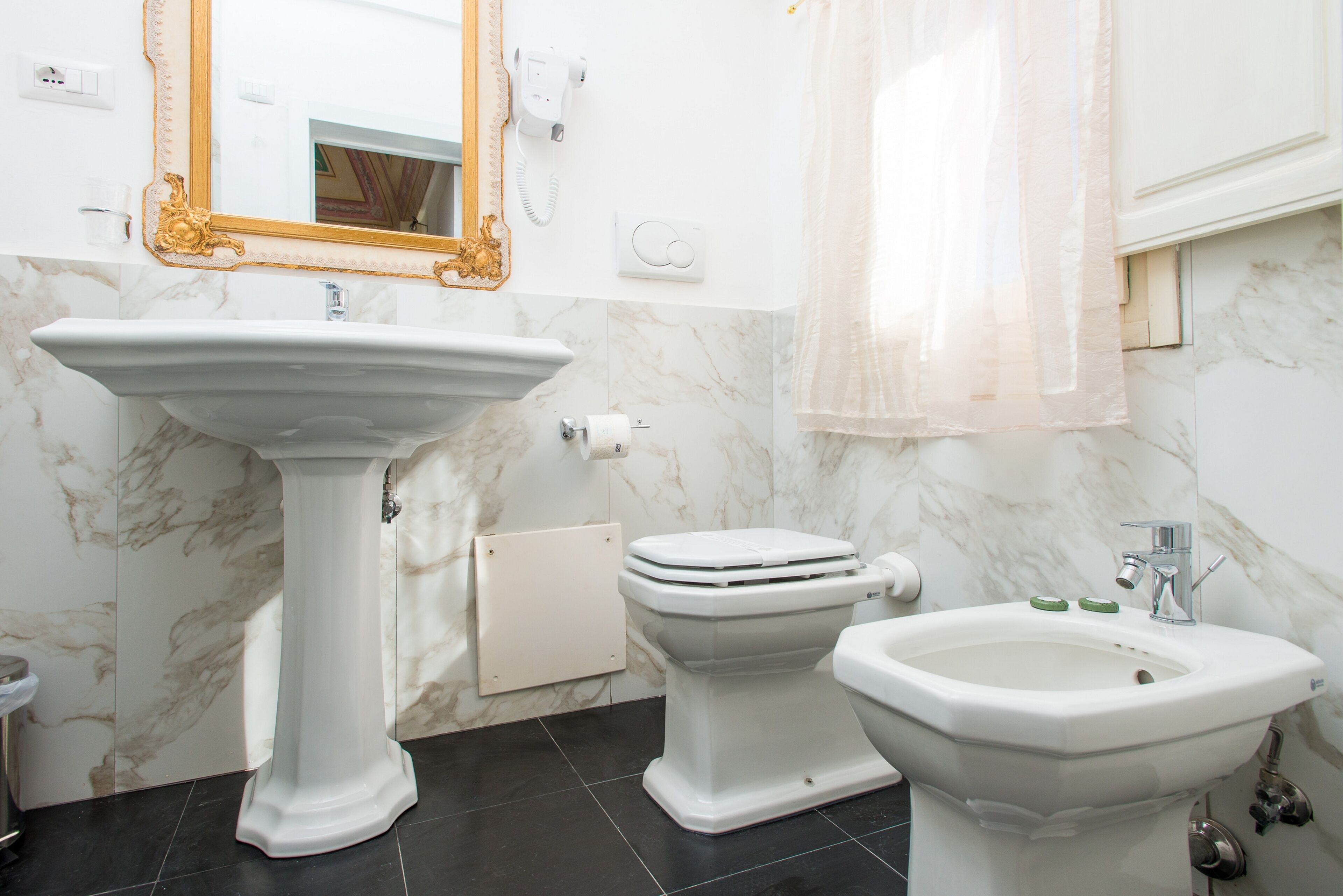 A bathroom with a pedestal sink, toilet, bidet, and a mirror with an ornate frame.