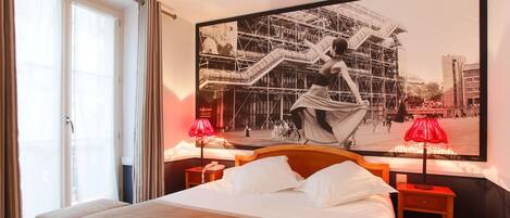 A hotel room with a large bed, a nightstand with a lamp, and a wall-mounted picture of a cityscape.
