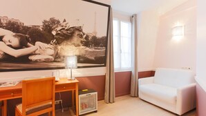 Quarto com uma grande obra de arte na parede, um sofá branco, uma escrivaninha de madeira com uma cadeira e um refrigerador pequeno.