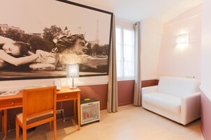 Una habitación con una obra de arte grande en la pared, un sofá blanco, un escritorio de madera con una silla y un refrigerador pequeño.