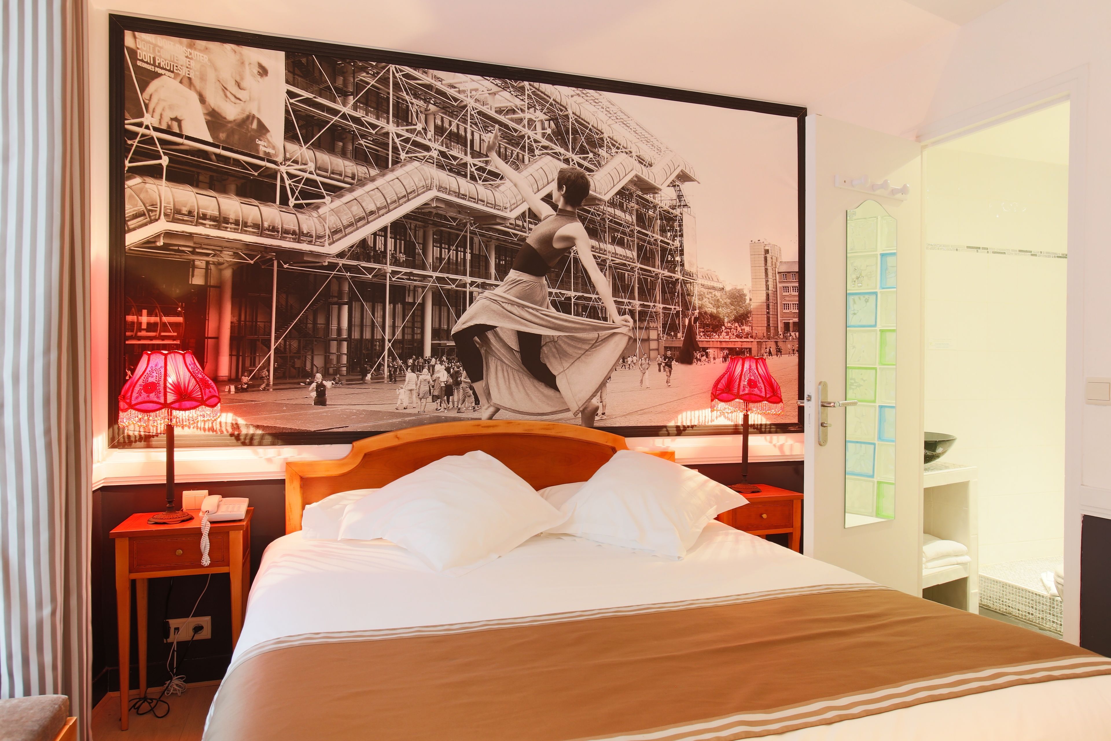 A hotel room with a large bed, two bedside tables, a nightstand, a red lamp, a large framed picture on the wall, and a window with a view of a building.