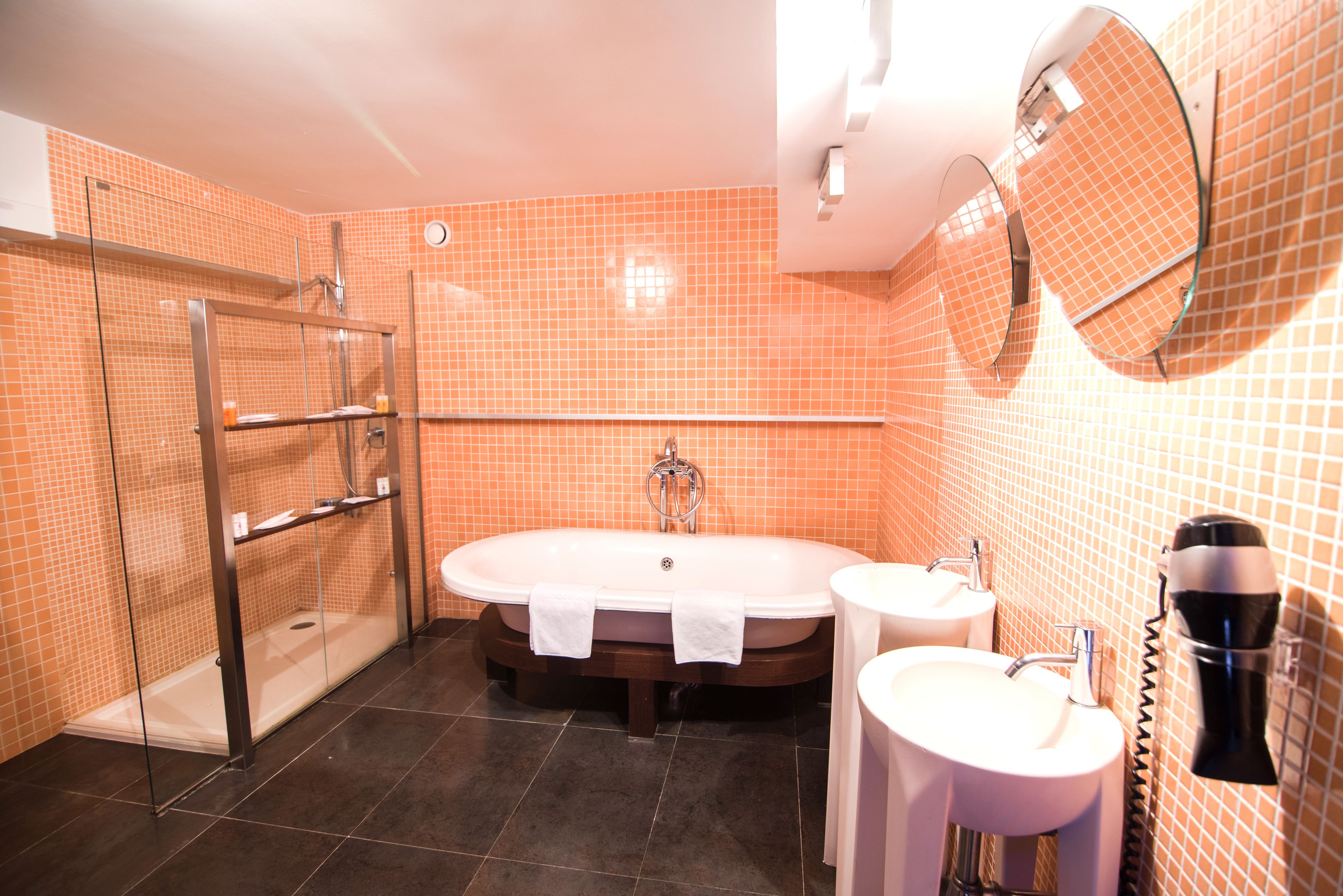 A bathroom with an orange tiled wall, a glass shower enclosure, a freestanding bathtub, two sinks, and a hairdryer mounted on the wall.