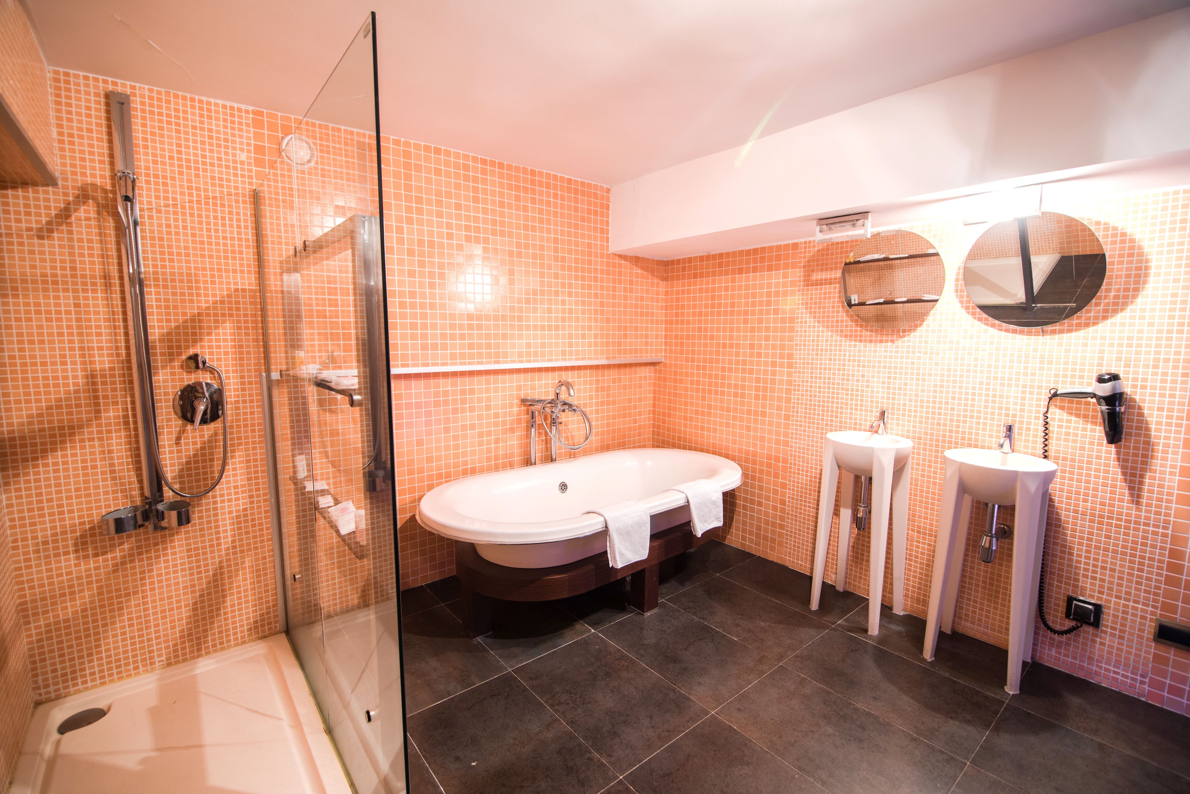 Een badkamer met een douche met glazen wand, een vrijstaand bad en twee wastafels op voet.
