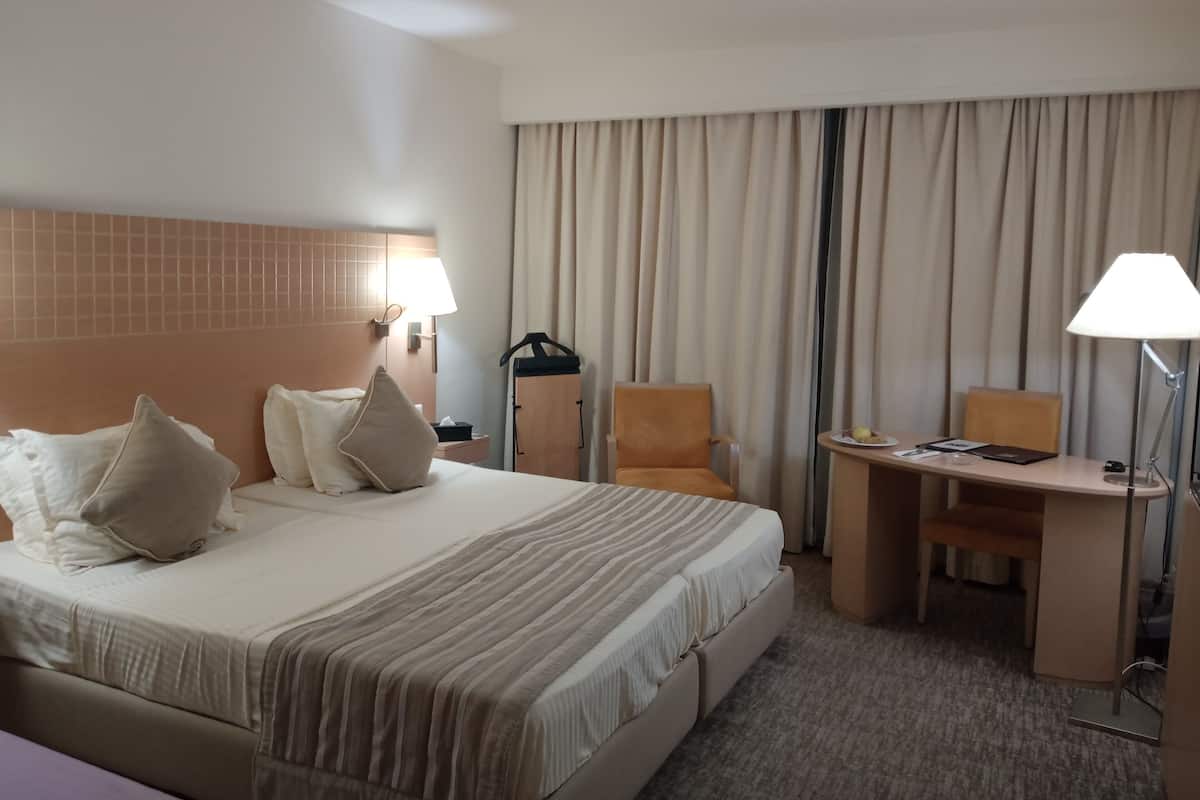 Quarto de hotel com uma cama grande, duas mesas de cabeceira com luminárias, uma escrivaninha com cadeira e uma janela ampla com cortinas.