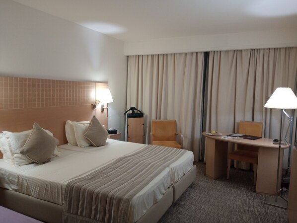 A hotel room with a large bed, two bedside tables with lamps, a desk with a chair, and a large window with curtains.