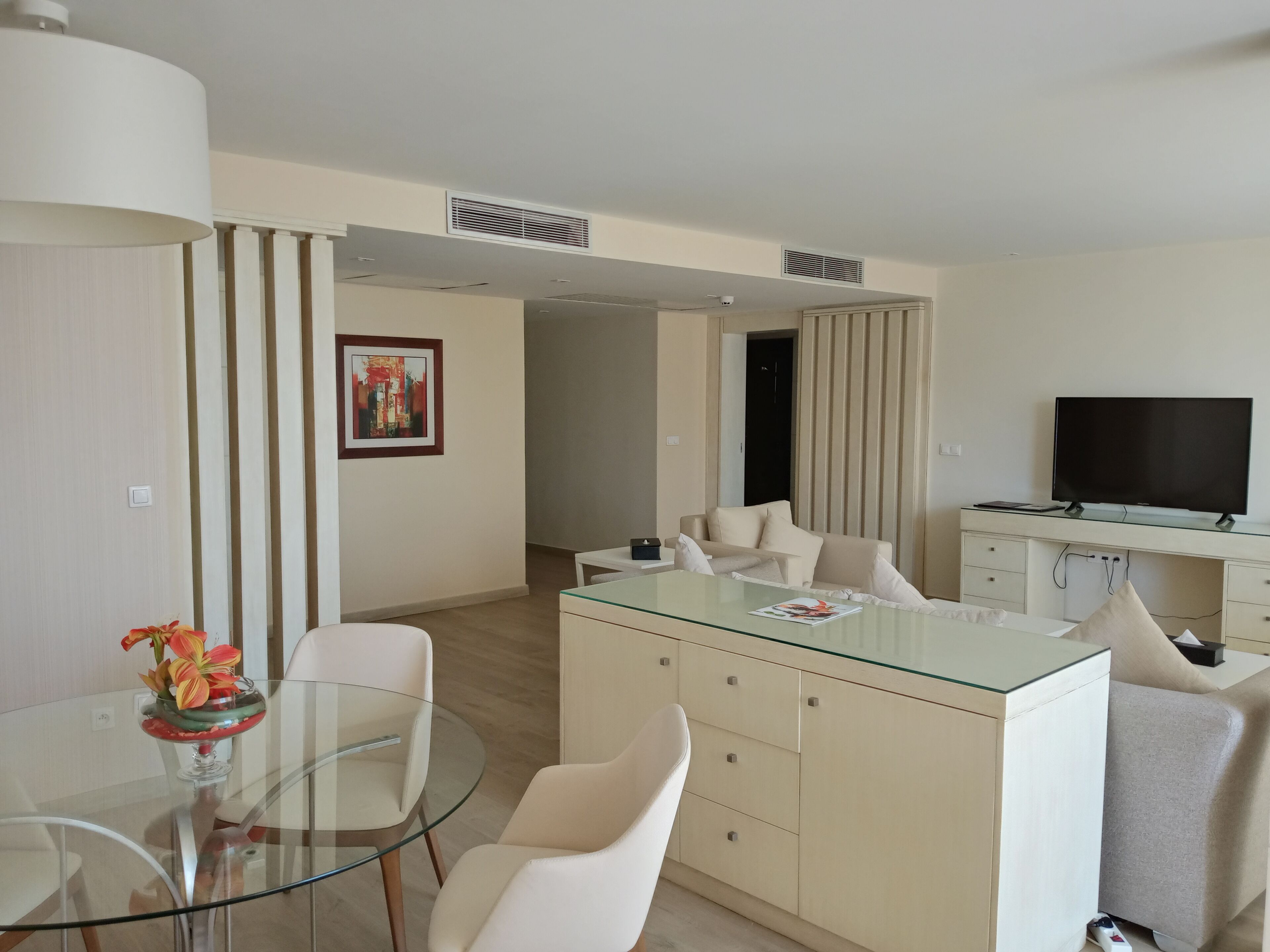 Un salón moderno con una mesa de comedor de cristal, sofás blancos, un televisor de pantalla plana y un jarrón decorativo.
