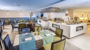 A modern restaurant with a central island, multiple dining tables, and a chef preparing food.