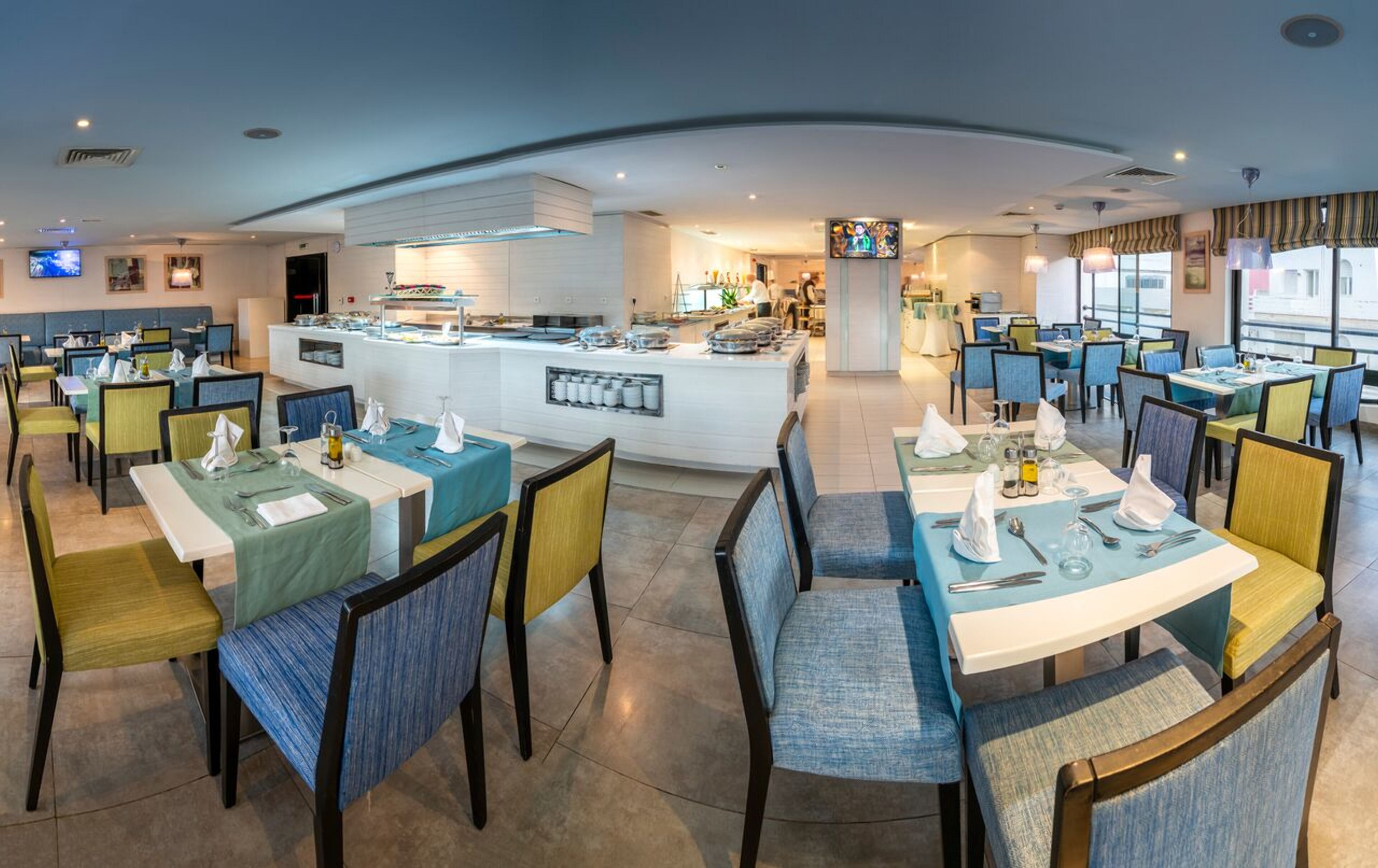 A modern restaurant with a central bar area, multiple dining tables with chairs, and a view of the kitchen in the background.