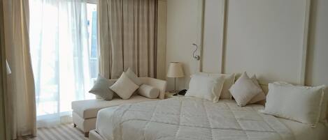 A hotel room with a large bed, a bedside table with a lamp, a chair, and a view of the city through sheer curtains.