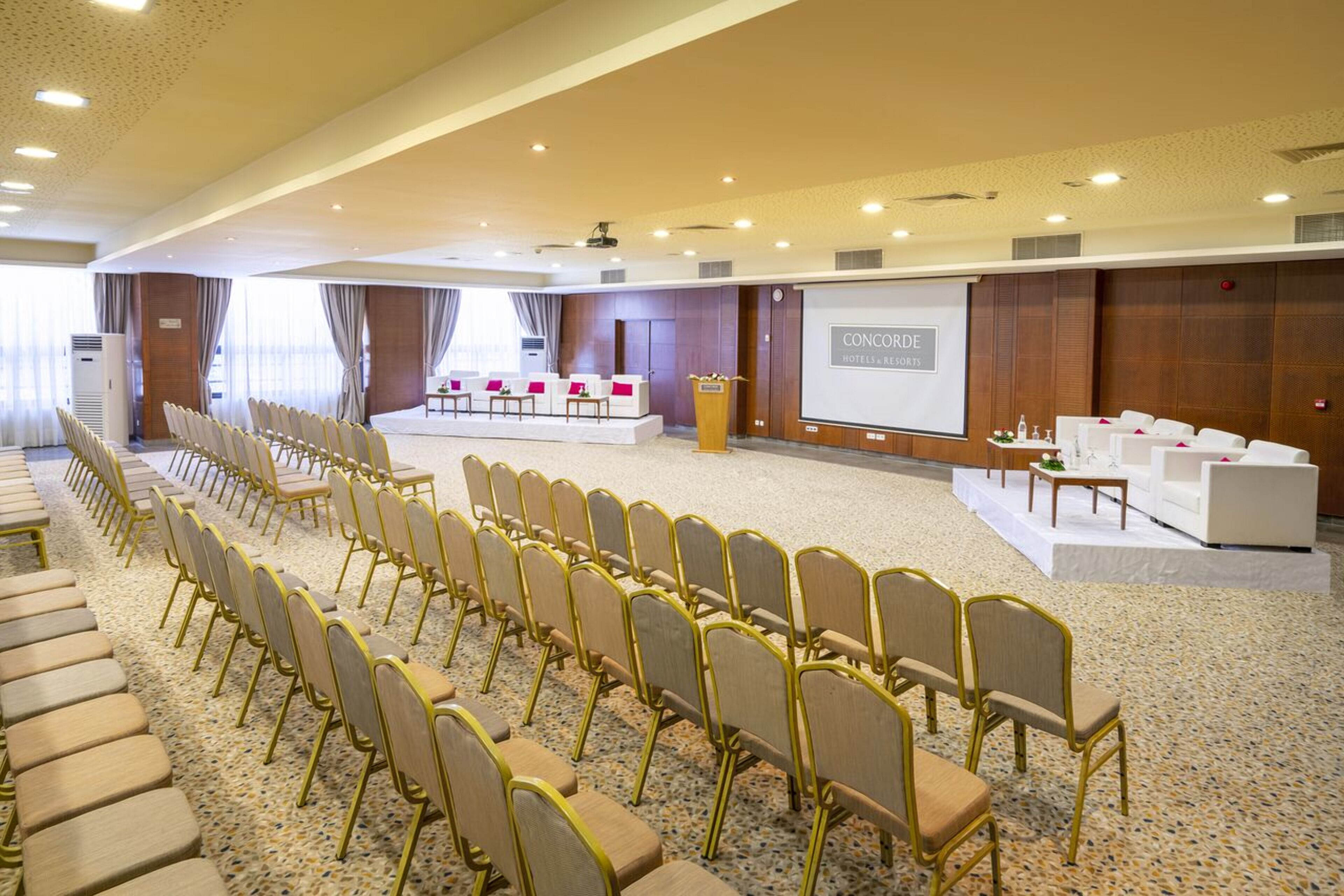 A conference room with rows of chairs, a stage, and a projection screen.