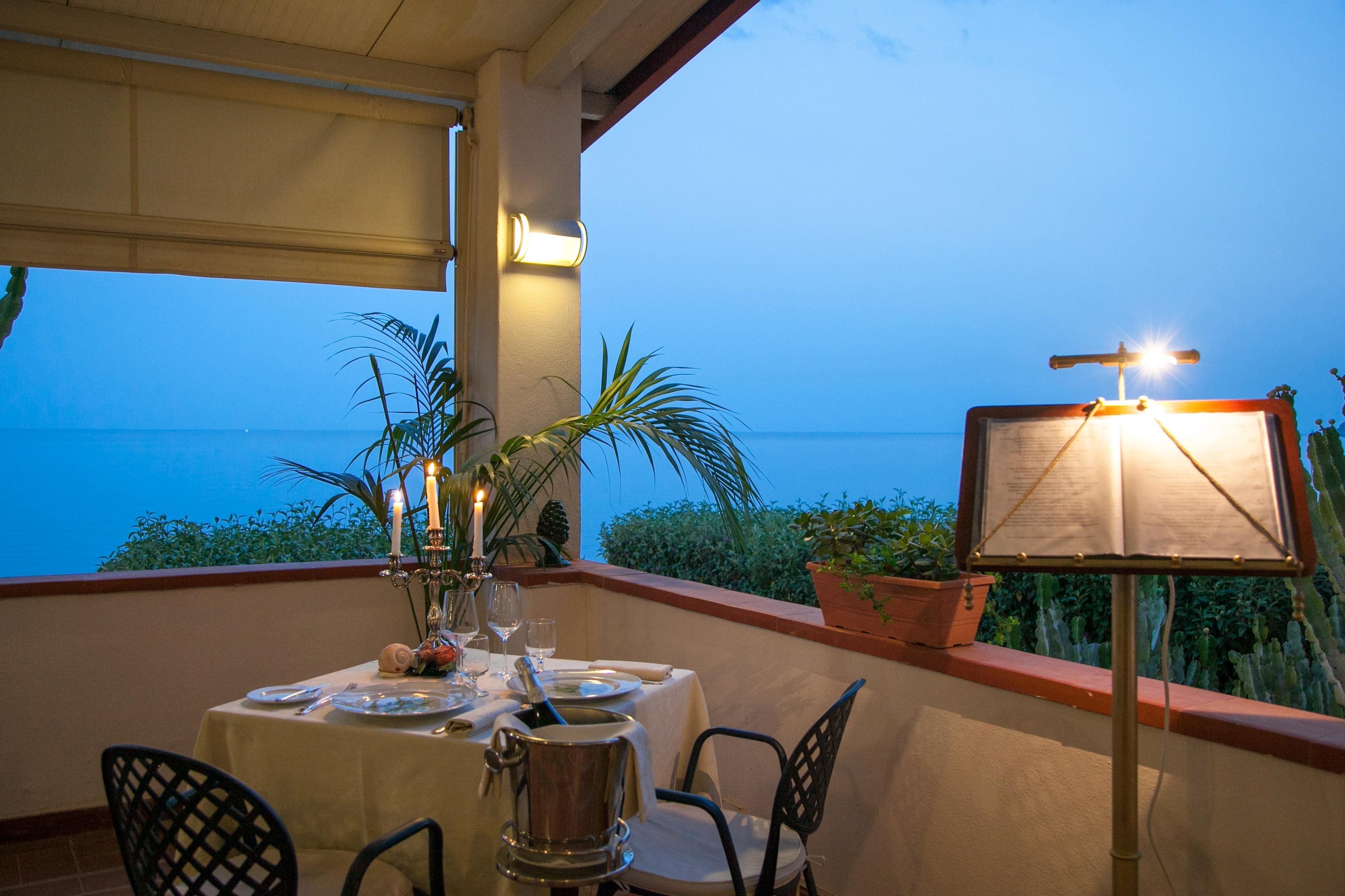A table set for dining on a balcony with a view of the sea, a candle centerpiece, and a menu stand.