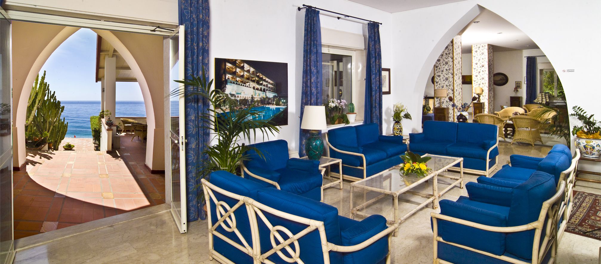 A spacious living room with blue furniture, a glass coffee table, and a view of the sea through an archway.