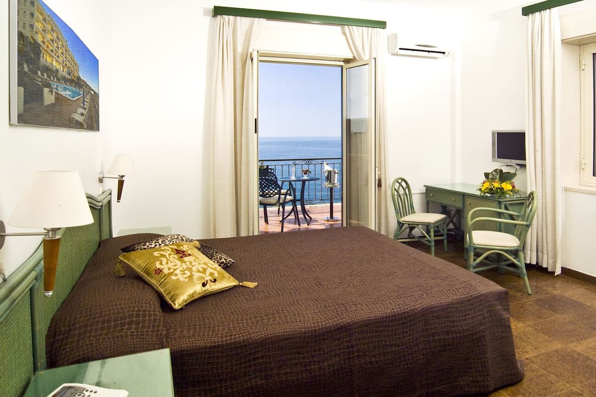 Ein Hotelzimmer mit einem Bett, einem Schreibtisch, einem Sessel, einem Fernseher, einem Balkon mit Meerblick und einem Bild an der Wand.