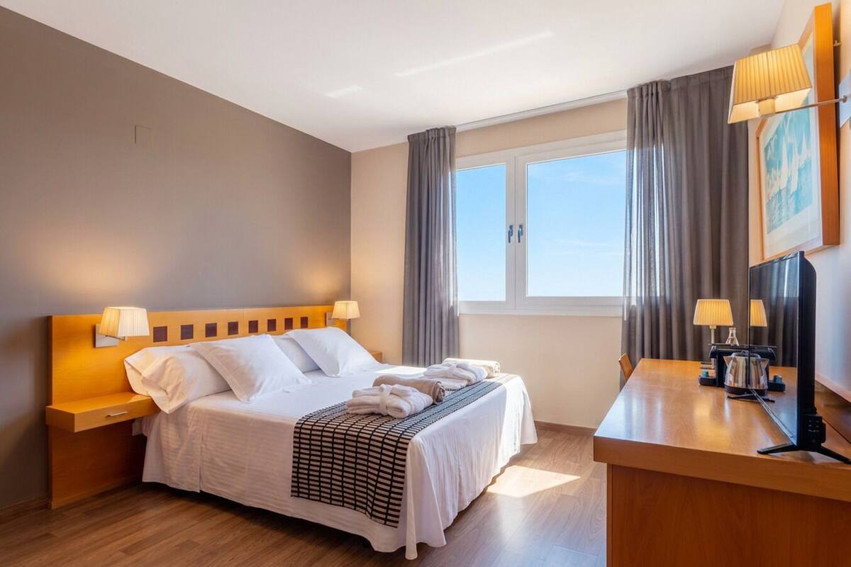 Habitación de hotel con cama, lámparas de noche, un escritorio con lámpara, ventana con cortinas y un cuadro en la pared.